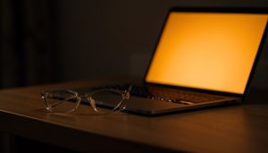 Clear eyeglasses on a wooden desk beside a laptop glowing with a warm amber screen in a dim room