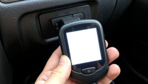 A handheld OBD2 code reader plugged into a car's diagnostic port with a lit screen
