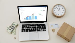An online seller's desk with a laptop showing a payments dashboard, cash, a bank card, a clock, and a shipping parcel