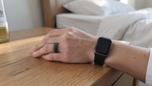 A hand wearing a smart ring and a smartwatch resting on a wooden nightstand by a bed